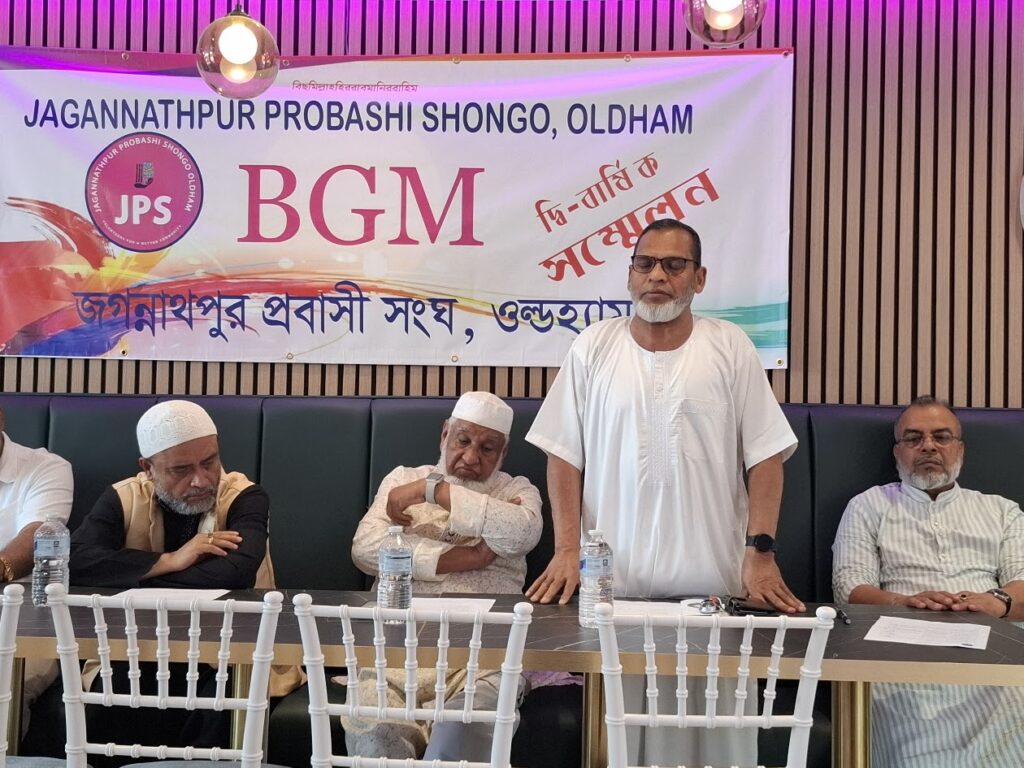 A member in white stands to speak at the Biennial General Meeting, with three colleagues seated at the table.